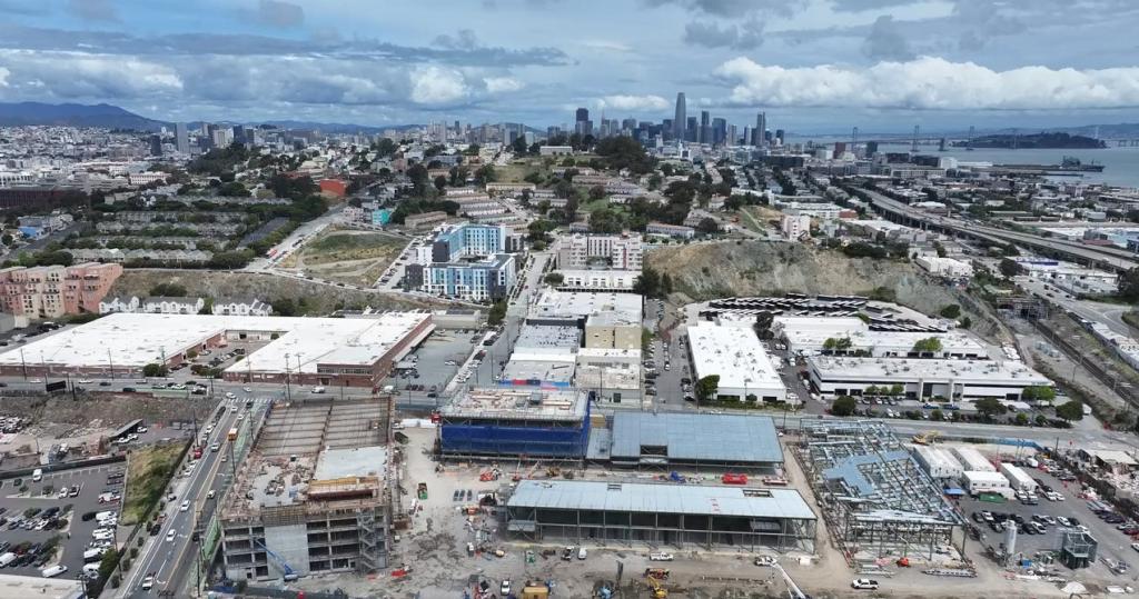 Vista aérea del campus que muestra el estacionamiento a la izquierda, el edificio administrativo y el almacén en el centro, el taller mecánico a la derecha y una vista de Potrero Hill con el perfil urbano del centro de la ciudad al fondo.