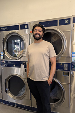 Smiling man with beard stands casually in front of stacked industrial dryers in a laundromat.