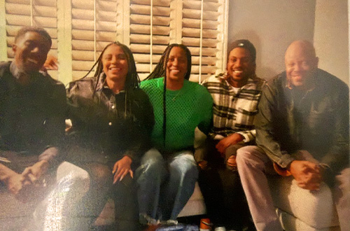 Five people sitting closely together on a couch in a warmly lit room with wooden blinds behind them.