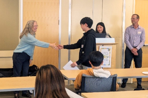 A woman with long hair shakes hands with a young man holding a certificate in a classroom setting.