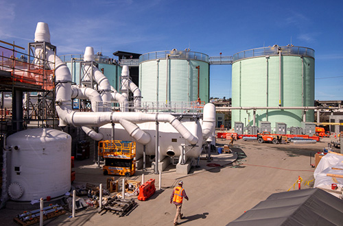 Industrial facility with large green storage tanks connected by extensive white piping under a clear blue sky.
