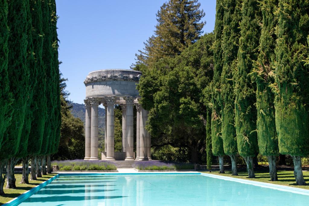 Image of the Pulgas Water Temple. The temple stands behind a bright blue pool of water lined by tall cypress trees.