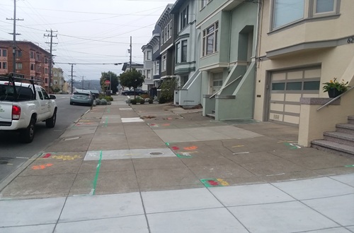 Wide sidewalk with concrete slabs, painted in places with colorful construction demarcations. No vegetation. Residential buildings to the right and a street with parked cars to the left.
