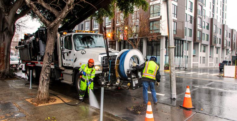 Các công nhân của SFPUC mặc đồ bảo hộ đang dọn dẹp vỉa hè thành phố gần một chiếc xe tải lớn chở thiết bị vào một ngày mưa.