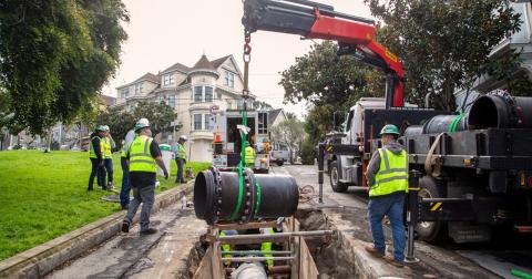Construction workers in safety gear install a large black pipeline using a crane over an open trench on a residential street.