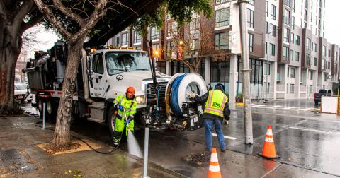 SFPUC workers in safety gear cleaning a city sidewalk near a large utility truck with equipment on a rainy day.
