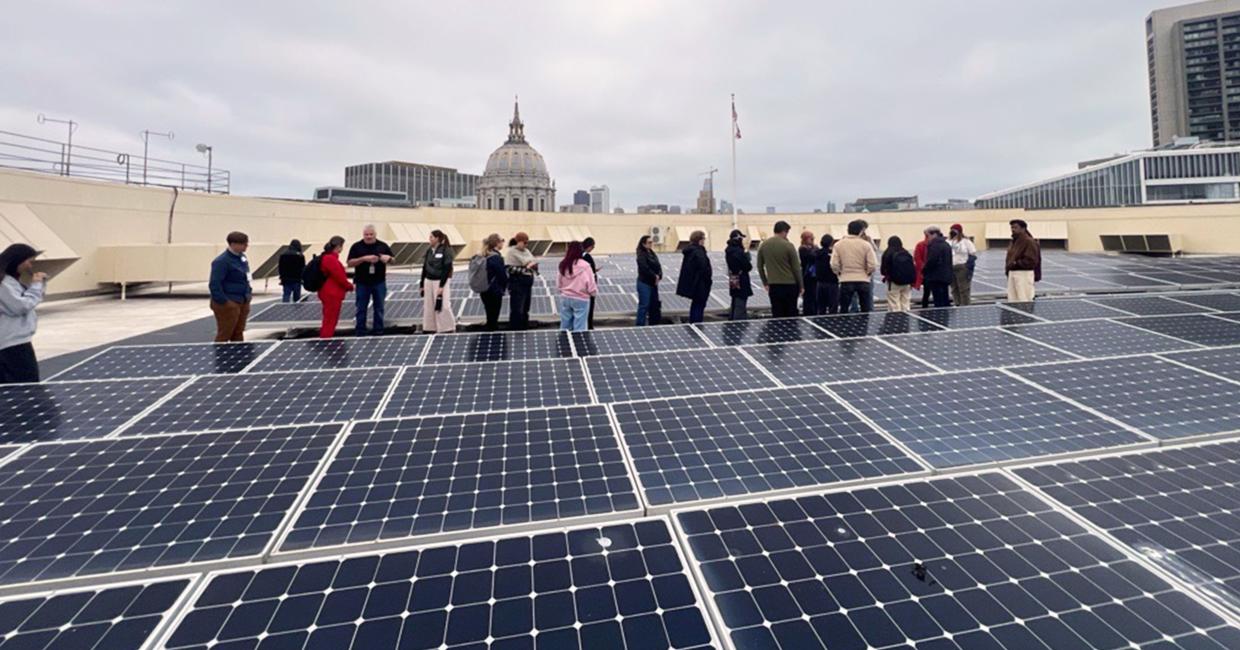 Group of people standing on a rooftop covered with solar panels, city skyline and dome building in the background under cloudy sky.