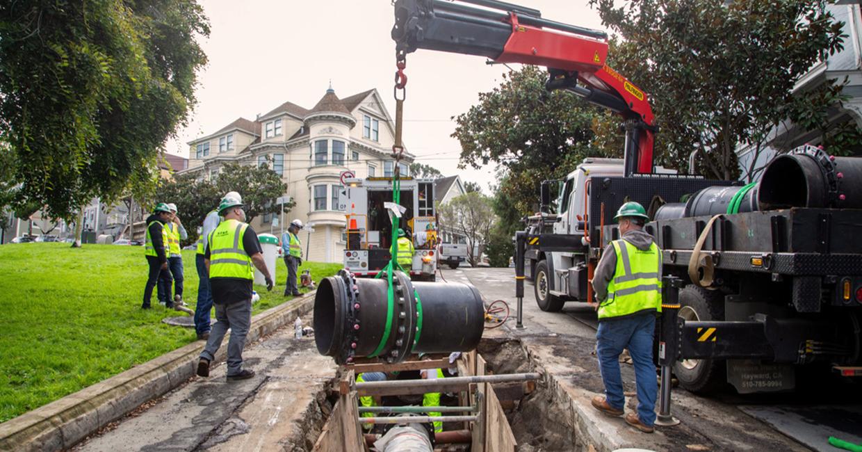 Construction workers in safety gear install a large black pipeline using a crane over an open trench on a residential street.