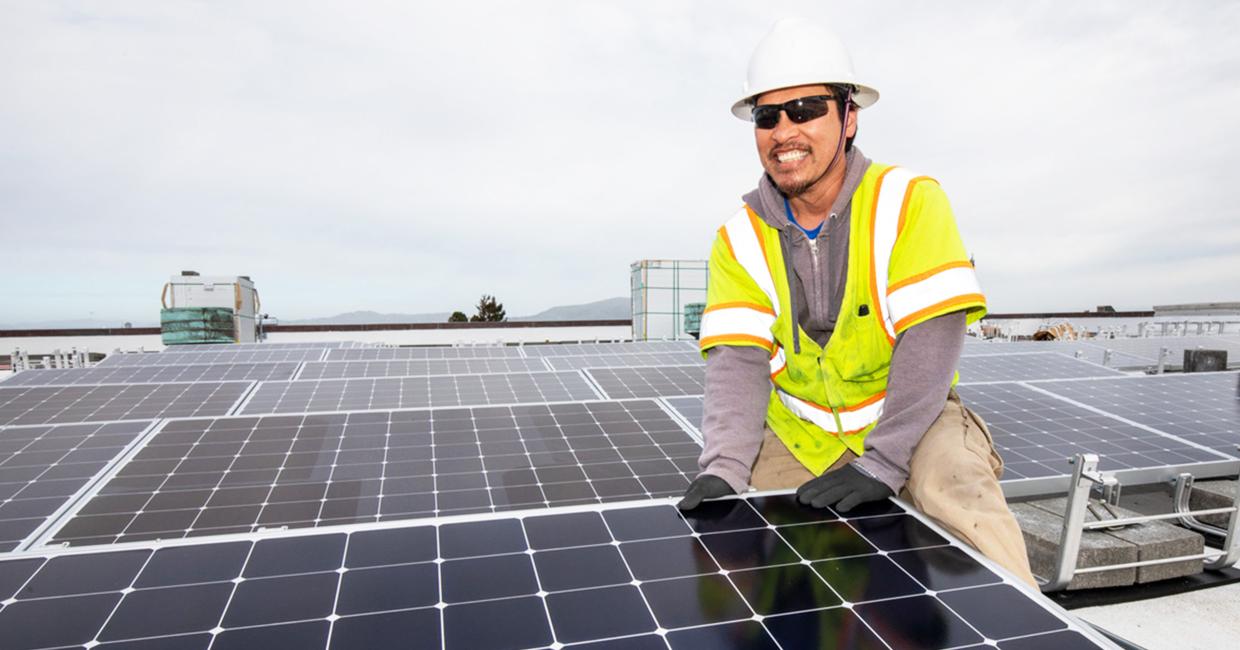 SFPUC Power Enterprise staff member in safety gear installing solar panels on a rooftop under a cloudy sky.