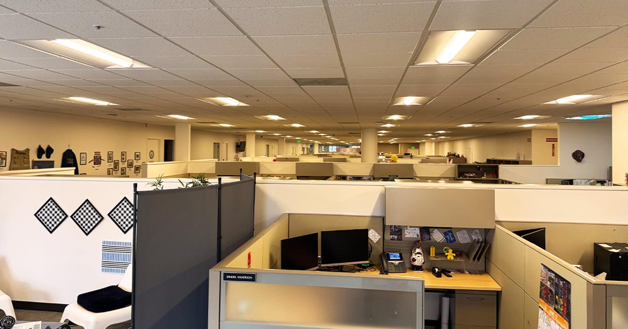 A spacious office with rows of cubicles, each equipped with computer monitors and personal items under modern LED lights.
