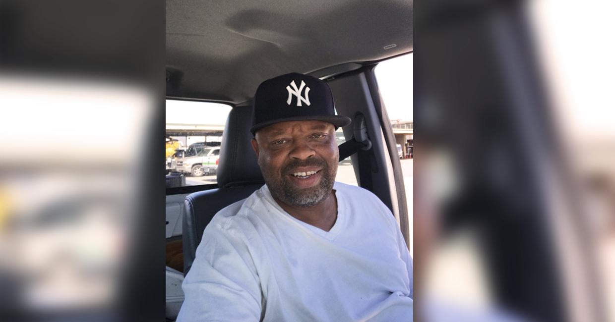 Man wearing a black New York Yankees cap and white shirt sitting in the driver's seat of a car with a parking lot visible outside.
