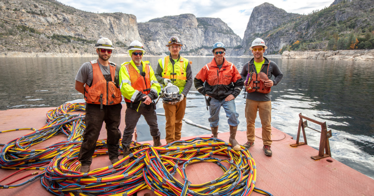 A team of commercial divers wearing safety vets and hard hats standing at a resrvoir.