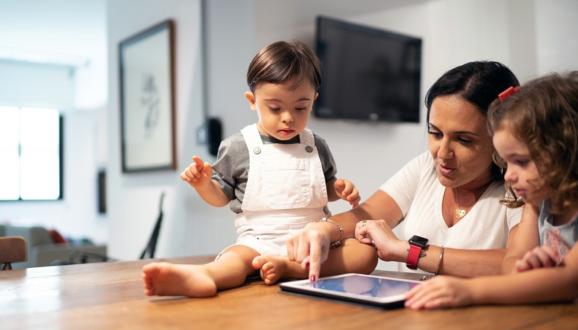 Mother and two children sit at a table looking at a tablet.