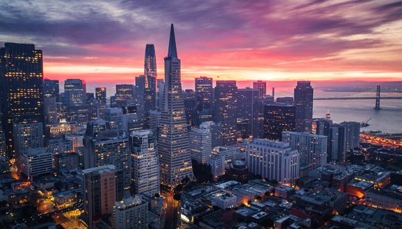 San Francisco skyline at sunset with the Transamerica Pyramid and Bay Bridge illuminated against a colorful sky.