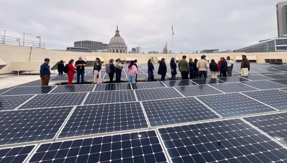 Group of people standing on a rooftop covered with solar panels, city skyline and dome building in the background under cloudy sky.