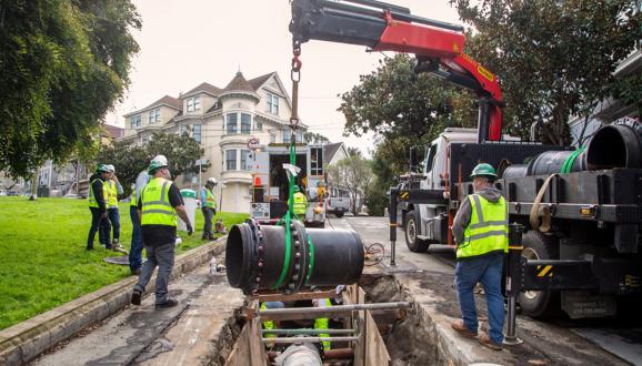 Construction workers in safety gear install a large black pipeline using a crane over an open trench on a residential street.