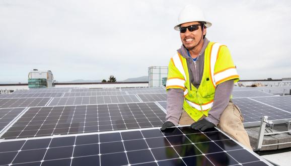 SFPUC Power Enterprise staff member in safety gear installing solar panels on a rooftop under a cloudy sky.