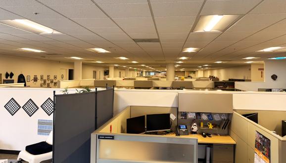 A spacious office with rows of cubicles, each equipped with computer monitors and personal items under modern LED lights.